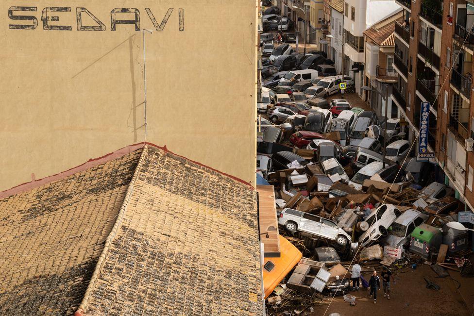 Aerial images of a building which has the word Sedaví painted on the wall. To the right a street is piled full of cars and debris as three people look at the damage 