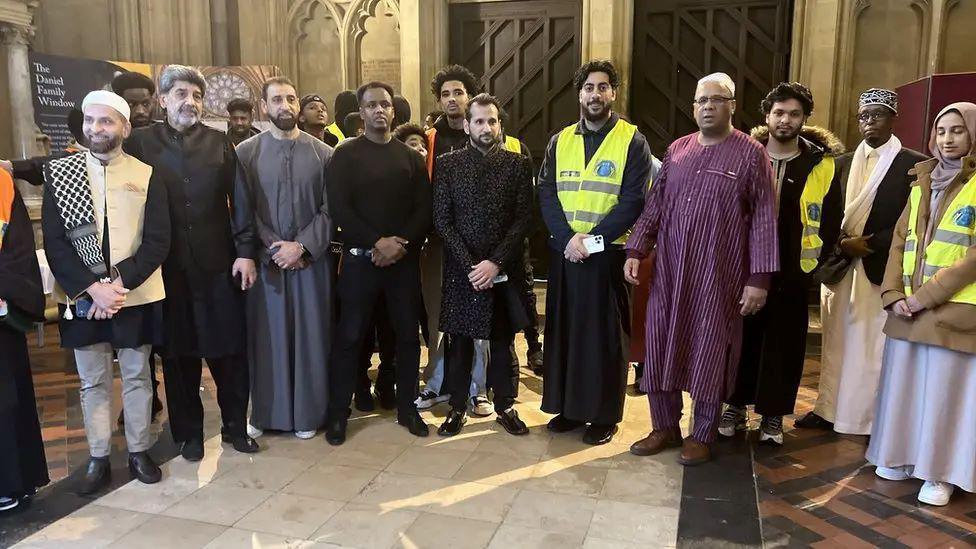 A group of men stand together in a group smiling at the camera. The men are dressed in religious dress for ramadan. The Cathedral walls are seen behind the group as they stand within the entrance. 