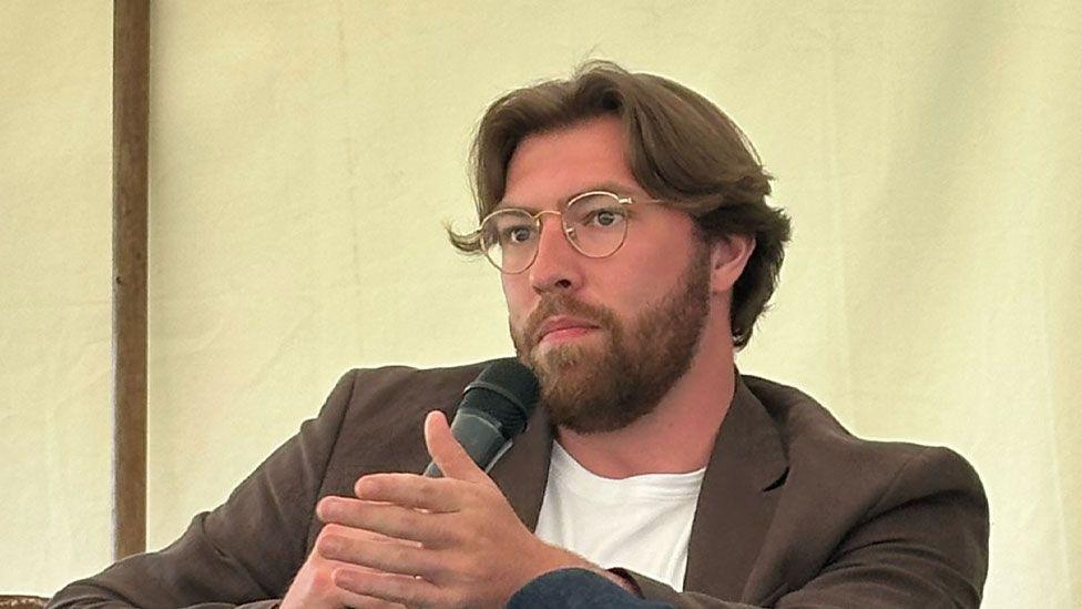 Jackson van Uden sitting on a stage at a history festival. He has wavy mid-brown hair and a brown beard and is wearing gold rimmed glasses and a brown jacket over a white T-shirt. He is looking to the left and is holding a microphone up to his mouth with his right hand while gesticulating with his left.