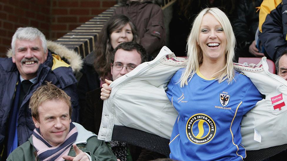 Soccer AM presenters Tim Lovejoy and Helen Chamberlain in a football stadium. He's seated, wearing a scarf and jacket. Helen is standing, with a beaming smile, holding open her jacket to reveal an AFC Wimbledon shirt with a Sports Interactive sponsor logo.