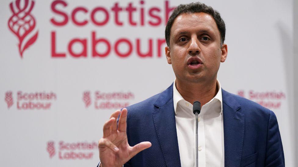 Anas Sarwar, with dark hair and wearing a blue suit with white shirt, speaks at a podium in front of a Scottish Labour sign 