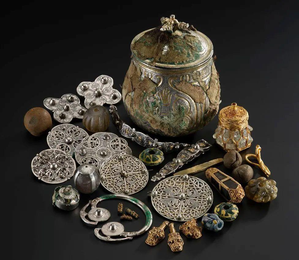 A collection of metal objects, including brooches and bracelets, and a metal vessel, displayed against a black background.