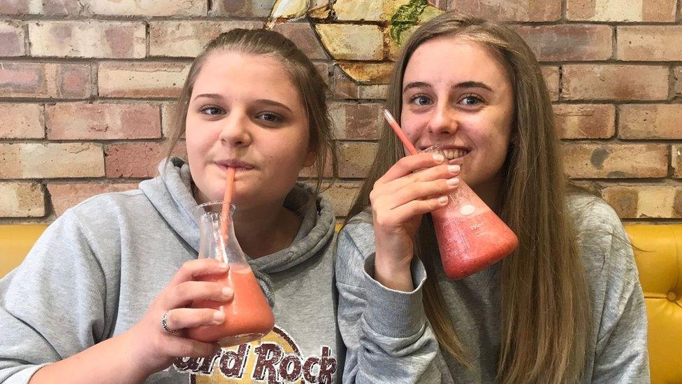 Gracie and Laura Nuttall sitting together, both drinking from a glass with a red-orange liquid in it that looks like a scientist's conical flash. Gracie is on the left and is wearing a grey Hard Rock Cafe hooded top while Laura is on the right and is wearing a long-sleeve grey jumper