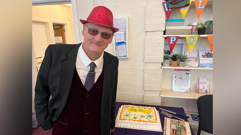 Pat poses for the camera next to his cake, which is a large white rectangular cake with orange and yellow decorations on and white icing. Pat wears a red sparkly hat, sunglasses, and a three-piece suit with blue tie and white shirt.