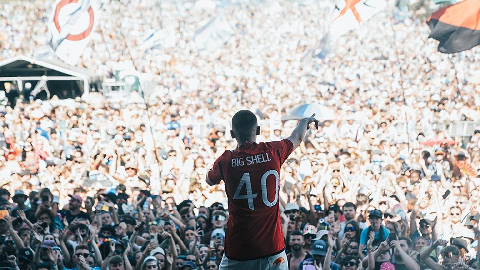 Aitch wearing the new Manchester United shirt, which is red. The back of the shirt says Big Shell with the number 40 below it. He is wearing white trousers, and is holding a microphone, with his right arm pointing at the crowd in front of him, full of thousands of people watching him perform. There are big flags in the crowd and many people on their phones filming his performance.