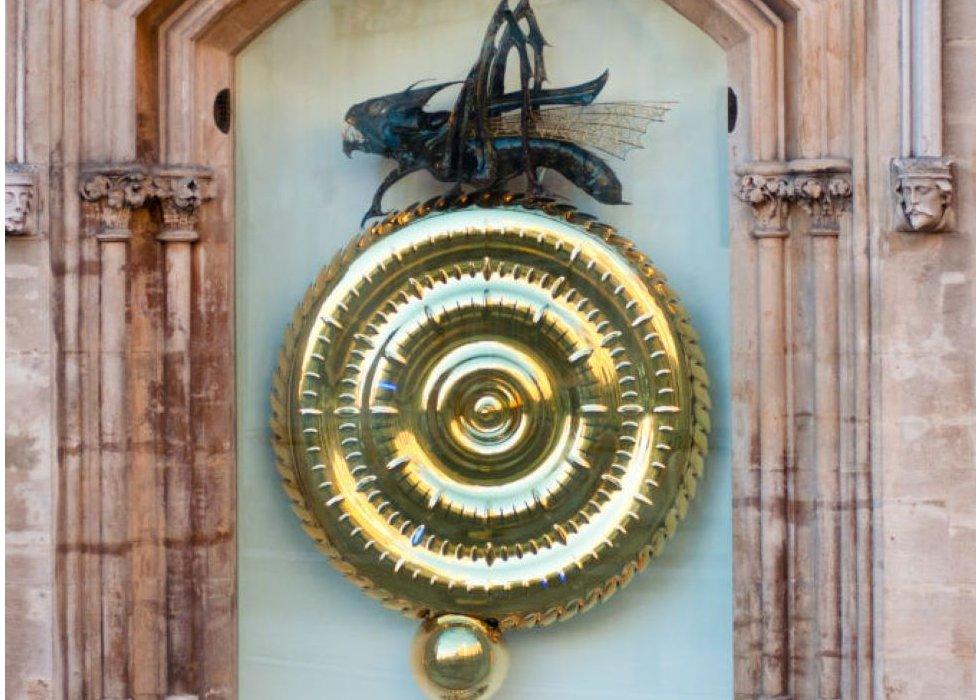 The clock in full shows a gold plated circle with a small gold sphere beneath it and a metal grasshopper sculpture sitting atop the large gold circle. The entire clock is in a corner of a building and protected by glass.