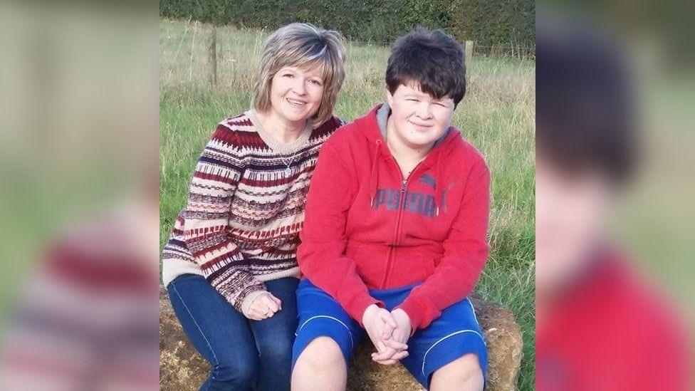 (L-R) Sharon wearing blue jeans and a purple and cream jumper, with Charlie who is wearing blue shorts and a red zipped hoodie both smiling at the camera.