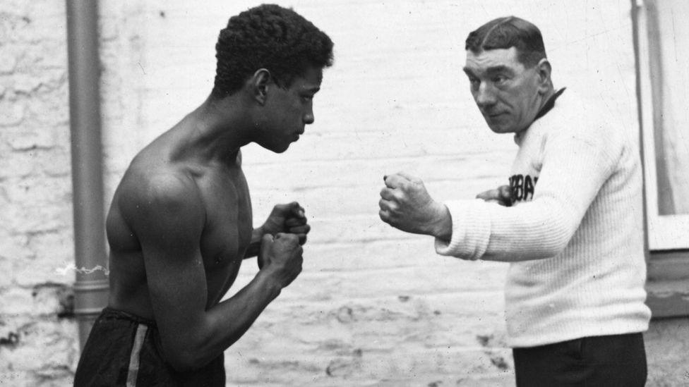 Len Johnson, wearing black shorts, sparring with his sparring partner, who wears a white jumper and black trousers