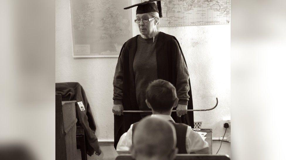 Black and white image of two young boys reenacting what it was like in a school classroom with their teacher during WWII
