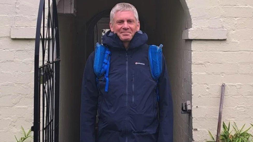 photo of Ian Evans. He is stood in a doorway wearing a navy blue coat. He is looking directly at the camera and smiling.