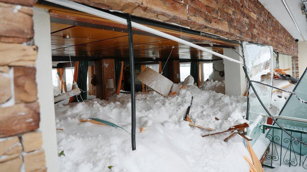 Snow is seen covering the inside room of a hotel, piled high enough to touch the ceiling lampshades, while the windows are shattered and broken