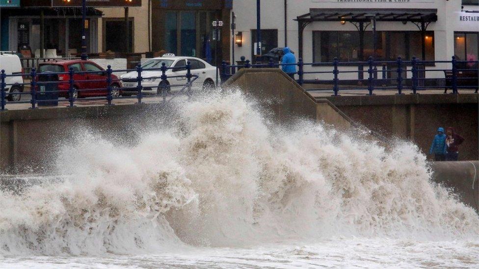 Pedestrians walk along the promenade as huge waves crash