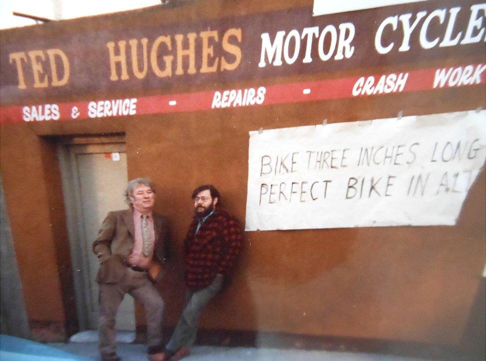 Seamus Heaney and Barrie Cooke posing under a sign for Ted Hughes motorcycles in Dublin (1985), just after Ted Hughes was appointed Poet Laureate