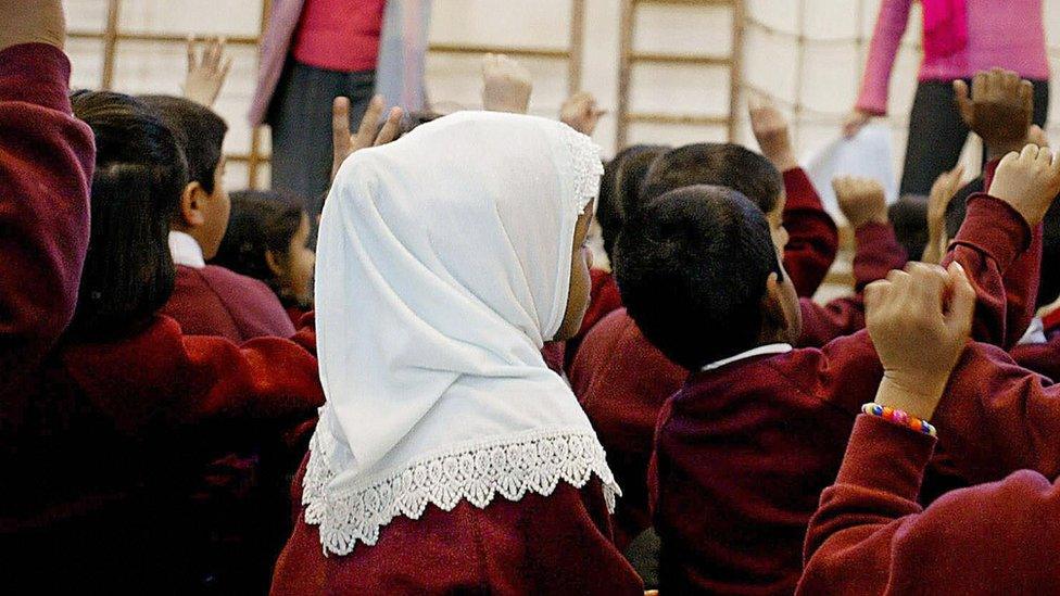 A young girl wearing a Muslim headscarf listens as two teachers instruct a class.