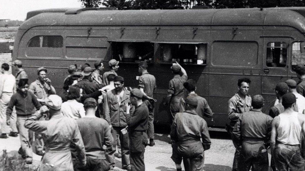 USAAF personnel receive food and drink from an American Red Cross Clubmobile. The vehicle is a bus which has two open flaps in its wall. Men are reaching up to the counter behind the window. Others are milling around, chatting and eating