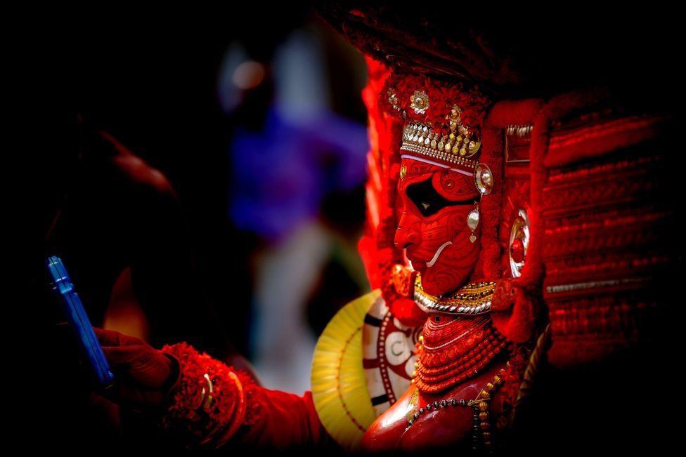 A performer in an elaborate red mask and headdress looking at their mobile phone