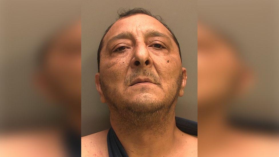 A police custody picture of a man with short black hair. He is wearing a black vest.