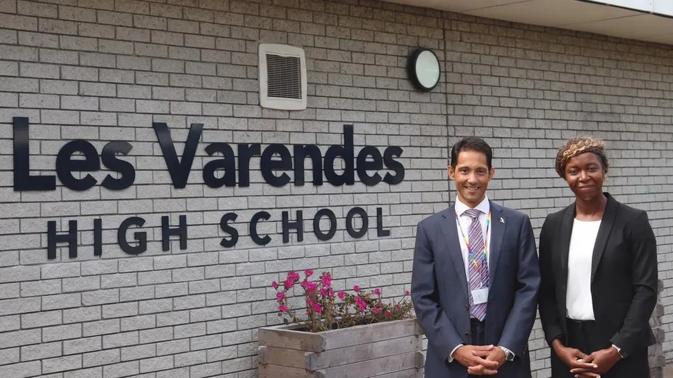 A man and woman in suits, Kieran James and Verona Tomlin, stand next to a sign for Les Varendes High School.