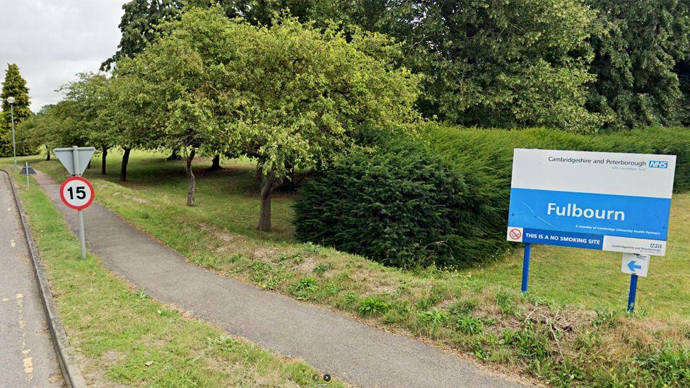 A sign at the entrance for Fulbourn Hospital, on the outskirts of Cambridge, on a sliproad next to a 15mph sign. In the background are trees.