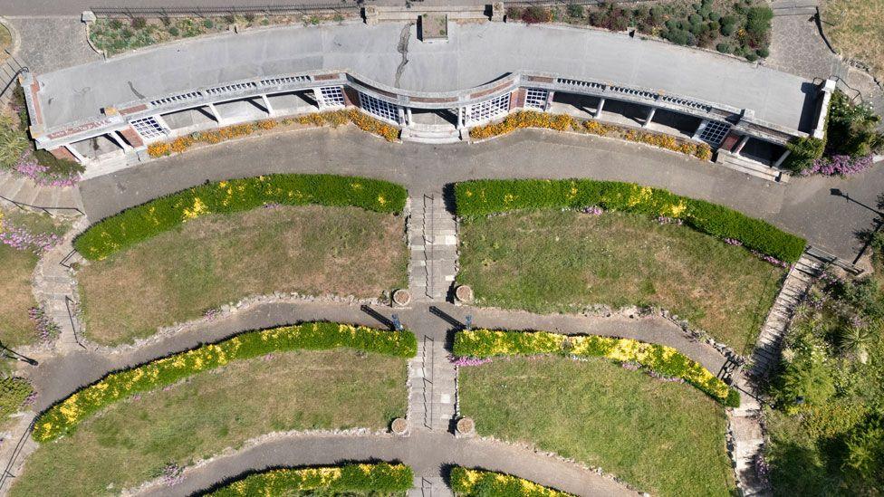An aerial shot of the Sun Shelter at Westcliff-on-Sea. At the top is the curving narrow single storey building. In front of it pavements and then three terraces of grass, edged by flower beds and bisected by pavements.