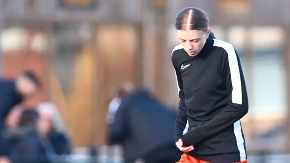 Picture of Sophia in football kit. She has dark hair tied in a ponytail and is wearing a dark shirt with a white stripe down the side