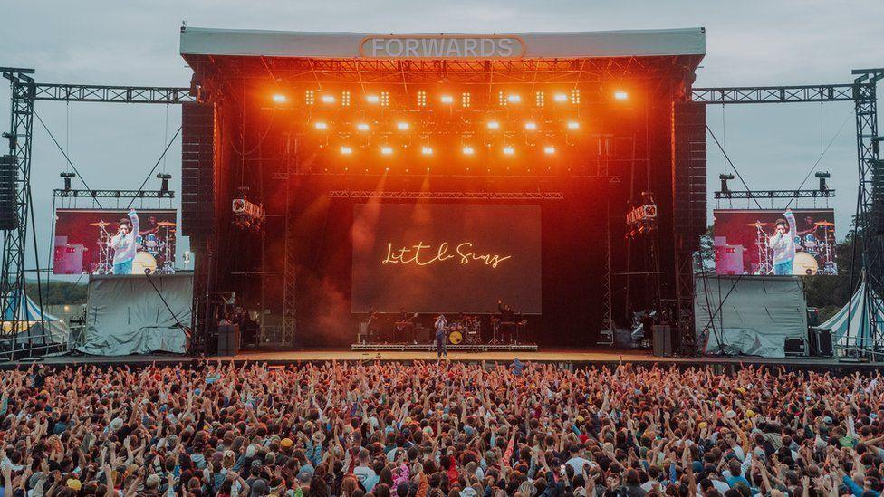 The Forwards Festival main stage lit up with orange lights at dusk in 2022. There is a large crowd of thousands of people with their arms up in the air. Little Simz is performing and her name is lit up in scrawling neon lights at the back of the stage. 