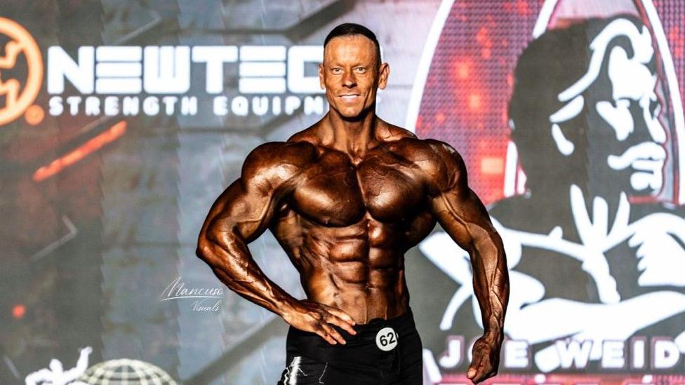 Mr Smith is posing on stage. He is muscly and tanned.