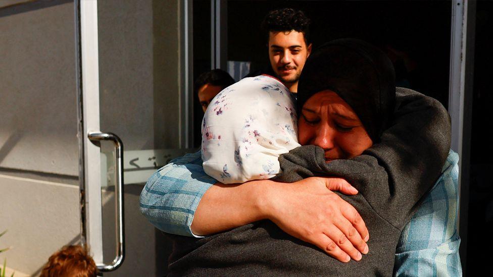 Two women, one with a white patterned headscarf and one with a black one, share an emotional embrace in a doorway. A young bearded man and another person - their face mostly obscured - look on in the background.