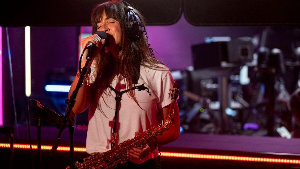 Abi Harding of The Zutons wears a white t-shirt and headphones and holds her saxophone as she sings in a purple and black radio studio