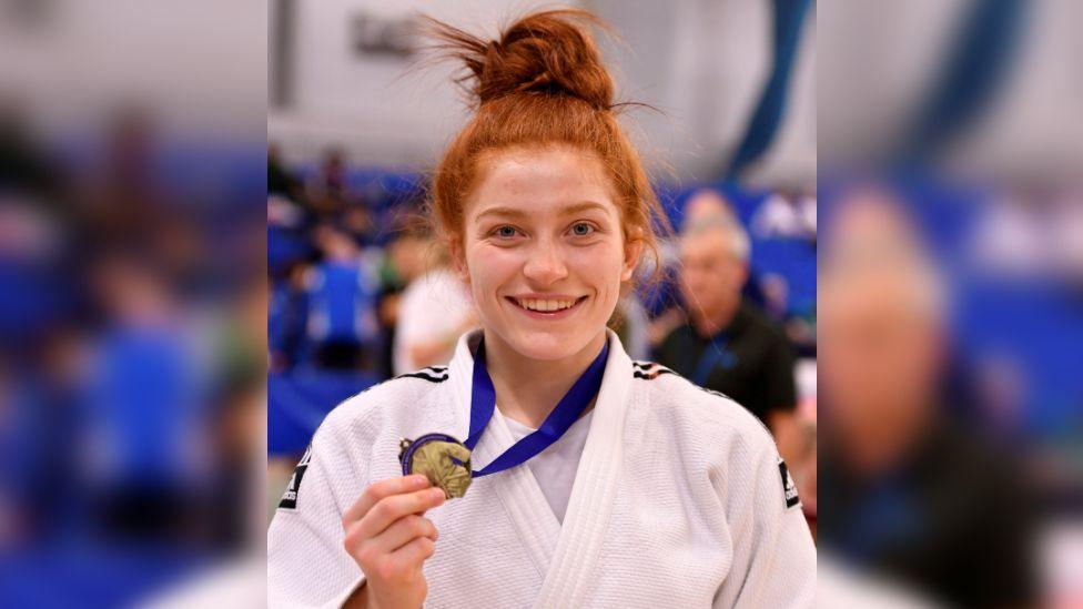 Lubjana Piovesana wearing her white judo uniform and holding out a medal with a blue ribbon round her neck. She has bright red hair which is tied up in a messy bun on top of her head. She is smiling at the camera, and behind her are blue stands for people to sit.