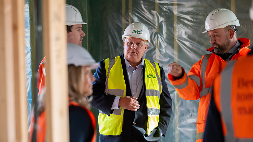 Three men and a woman in hard hats and high-vis jackets on a building site discussing things