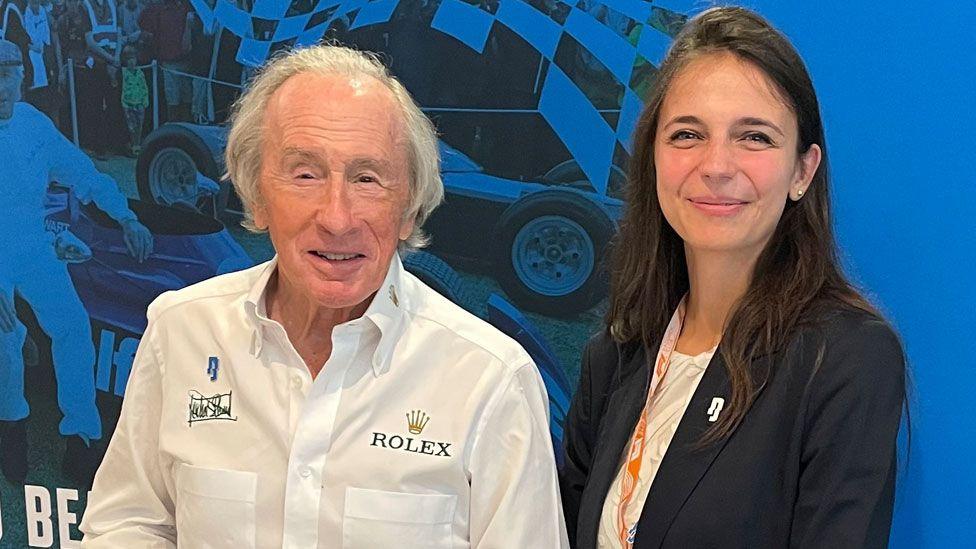 Sir Jackie Stewart and Dr Maura Malpetti, both smiling.  Sir Jackie is on the left and has ear-length white hair, swept back from his forehead, and is wearing a white shirt. On the right, Dr Malpetti has dark long hair over her shoulders. She is wearing a blue jacket over a white top.