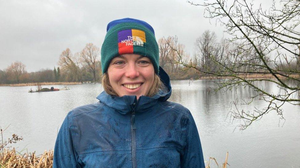 Smiling woman with fair hair wearing an anorak and a blue and green woolly hat stands next to a river