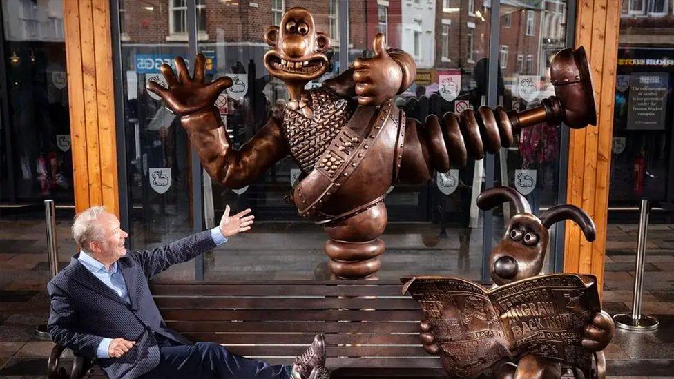 Nick Park sits on the Wallace and Gromit bench outside Preston market. He is wearing a blue suit and has his feet up and is pointing to Wallace in the Wrong Trousers behind him. Gromit sits at his feet reading the paper in the bronze-coloured sculpture