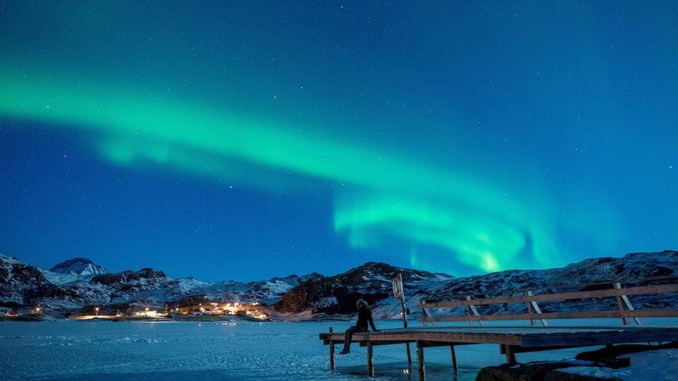 Northern lights over snowy mountains with a person sitting on a pier