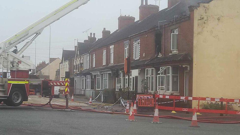 The front of the house with a fire engine parked outside showing fire damage to an upstairs window and the roof. The area around the house has been cordoned off with orange and white barriers