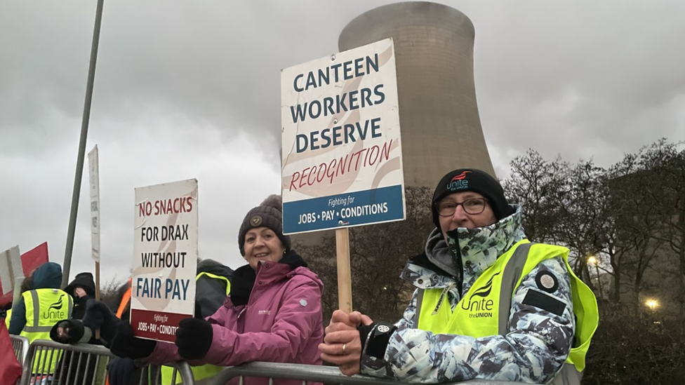 Workers stand on the picket line outside Drax power station. They're are wearing high-vis and holding placards. A cooling tower can be seen in the background.