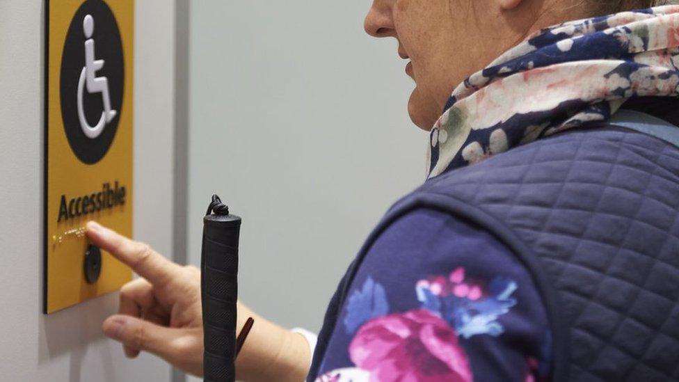 A visually impaired woman with a stick reads the braille on a disabled toilet sign.