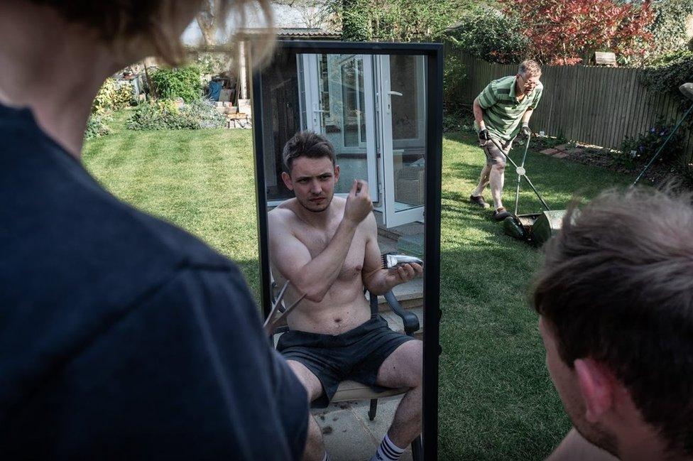 Man cuts his own hair in mirror as garden as other man mows the lawn