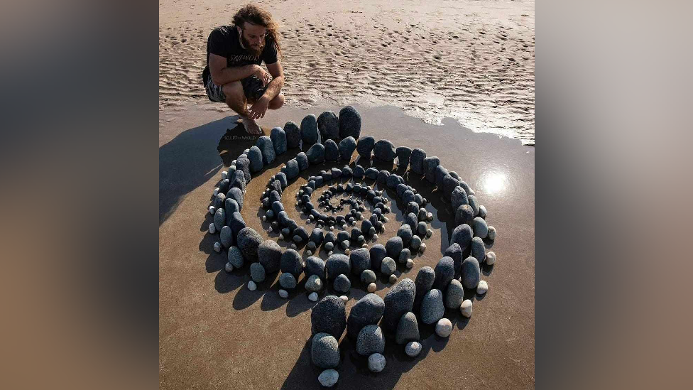 Man crouched down on the sand of the beach with a collection of stones carefully placed in a spiral formation. 