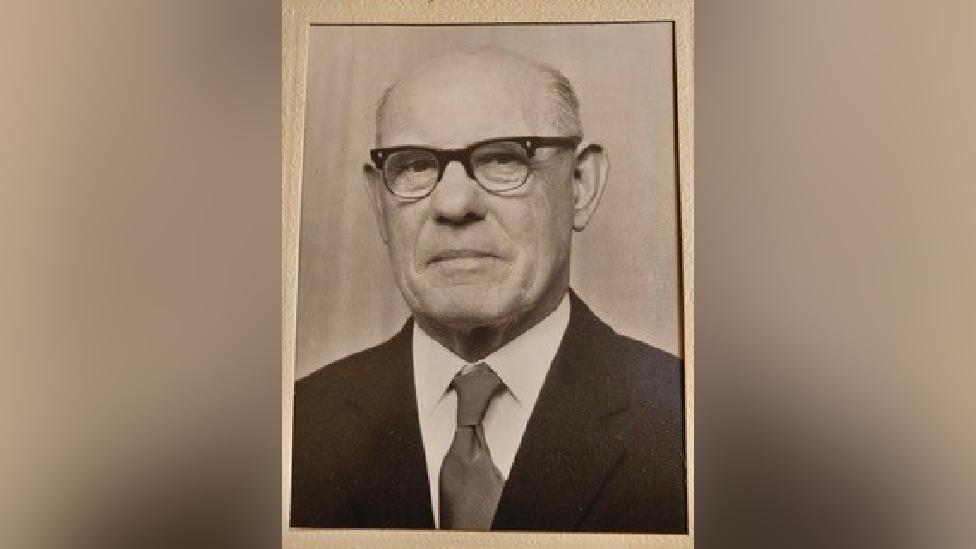 Ness Edwards, a man with black rimmed glasses, slightly frowning at the camera. He is wearing a black suit with a white shirt and a dark-coloured tie