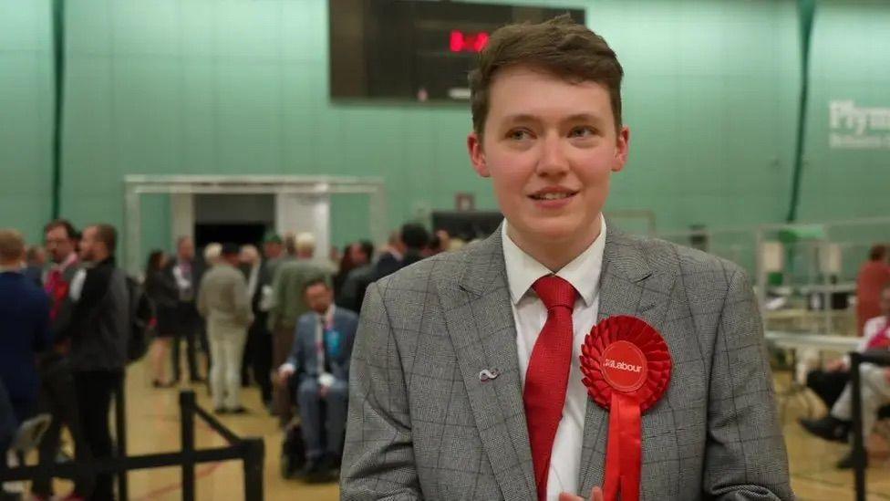 Dylan Tippetts in a white shirt, red tie and grey check suit with a Labour rosette