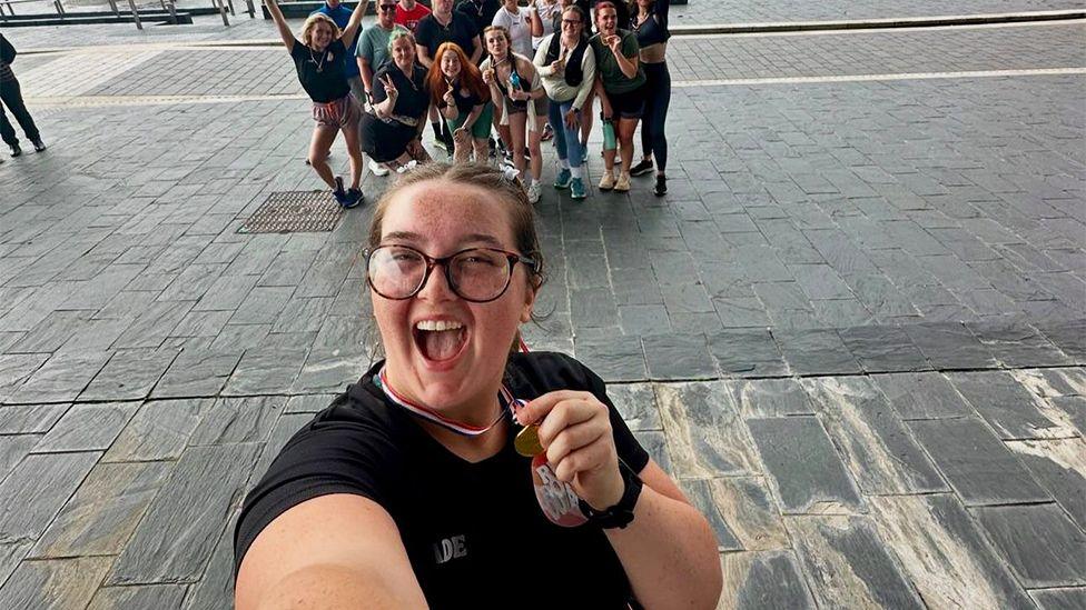 Jade taking a selfie while posing with a running medal. Jade is wearing a black top, dark rimmed glasses and has an open-mouthed expression. In the photo she has also included a number of fellow runners behind her.