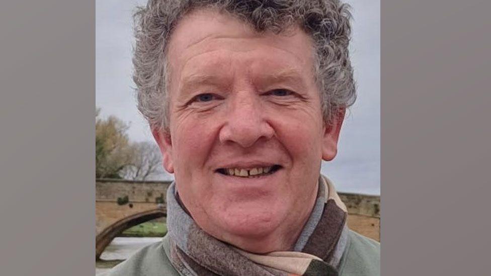 Neil Harvey standing outside with a glimpse of a river and bridge behind him. He has short curling salt-and-pepper hair, has a scarf wrapped round his neck and is smiling broadly