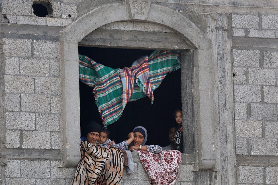 Women and children peer out of the window of a stone building. There are rugs draped over the window and hanging from the ceiling.