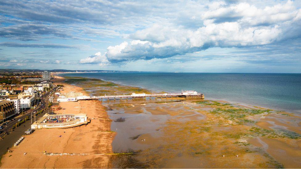 A drone shot of Worthing Pier. The tide is out and the beach can be seen next a number of buildings. The sea is blue and the low tide area is a yellow/brown colour. 