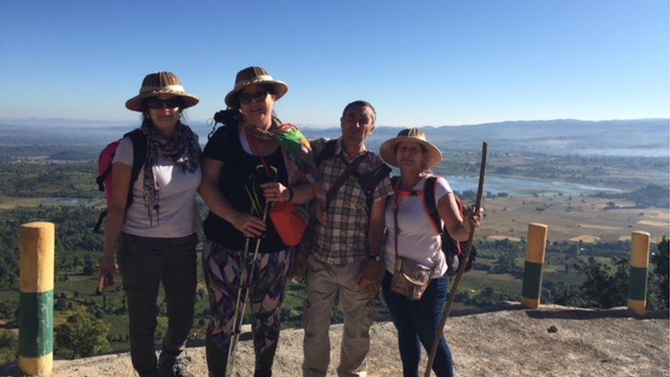 Four people stand in hot-weather clothing in front of a picaresque view of Myanmar