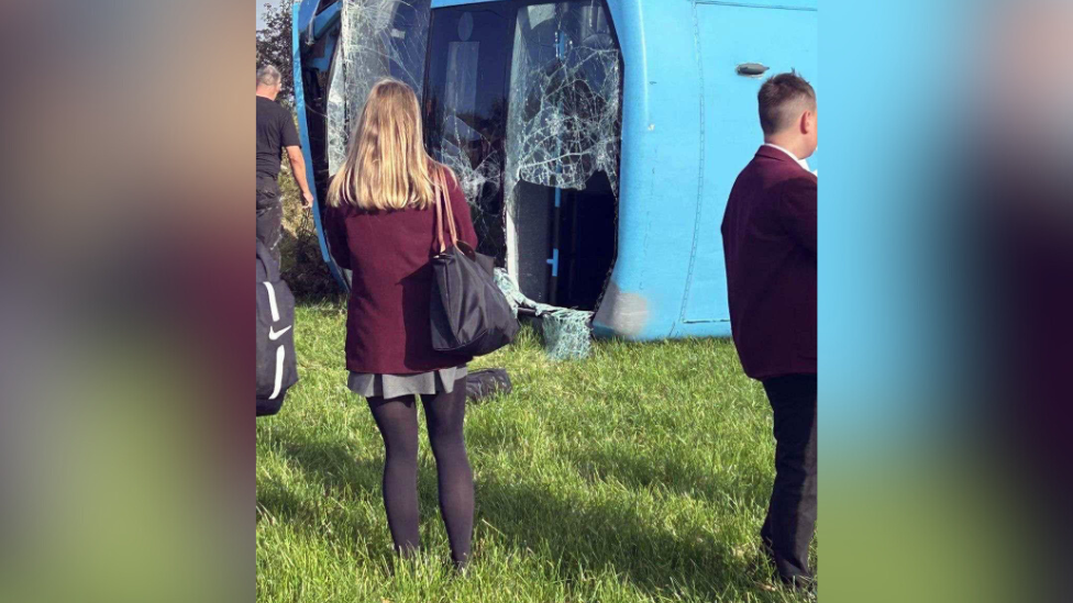 A still of a overturned blue, double-decker bus in a field. It has smashed windows while a pupil in a maroon blazer, grey skirt and dark tights looks on.