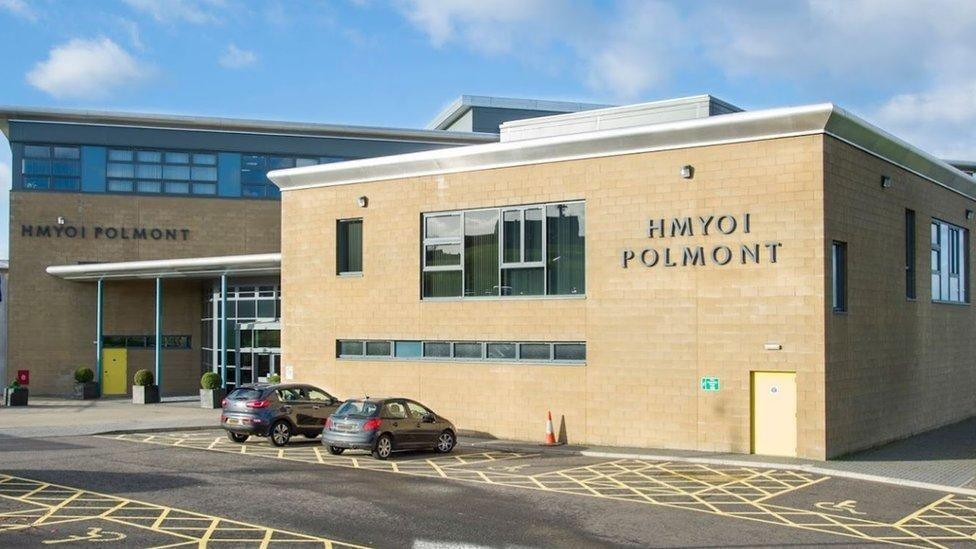 The outside of Polmont Young Offenders Institution. There appear to be two buildings which are both sandy-coloured and have HMYOI POLMONT letters on them. There is an entrance between the buildings and two cars parked outside in disabled spaces. The spaces are designated by yellow lines and the sign of someone in a wheelchair.