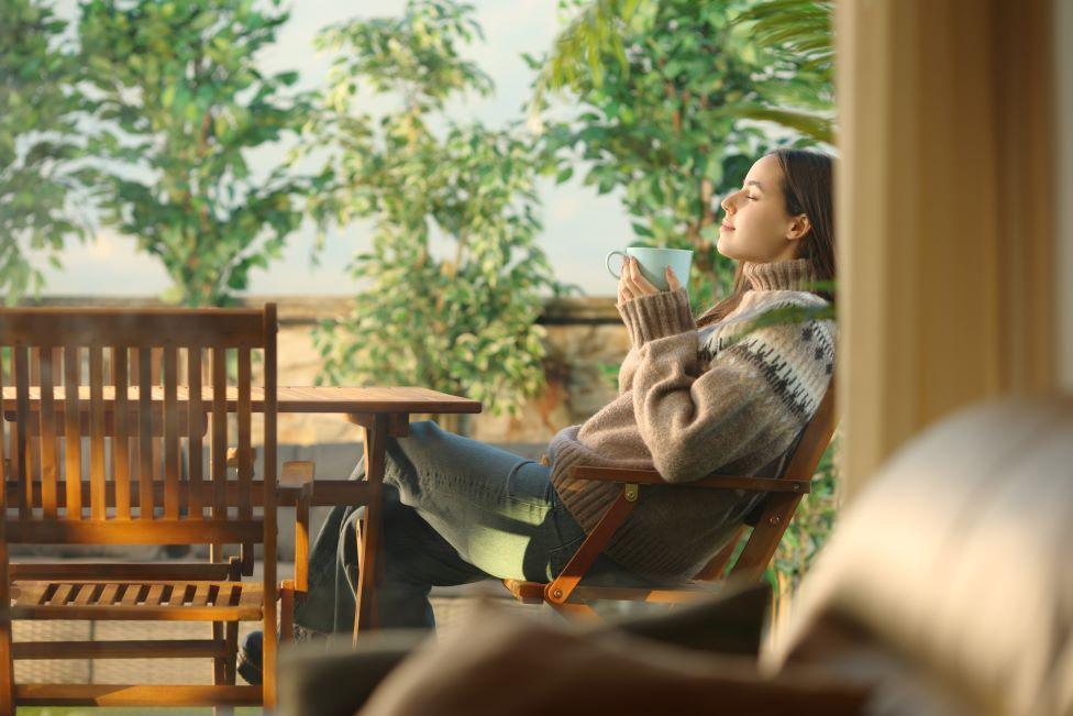 A woman in a jumper sits on a terrace outside a house, holding drink in a mug and sitting back with a contented expression on her face